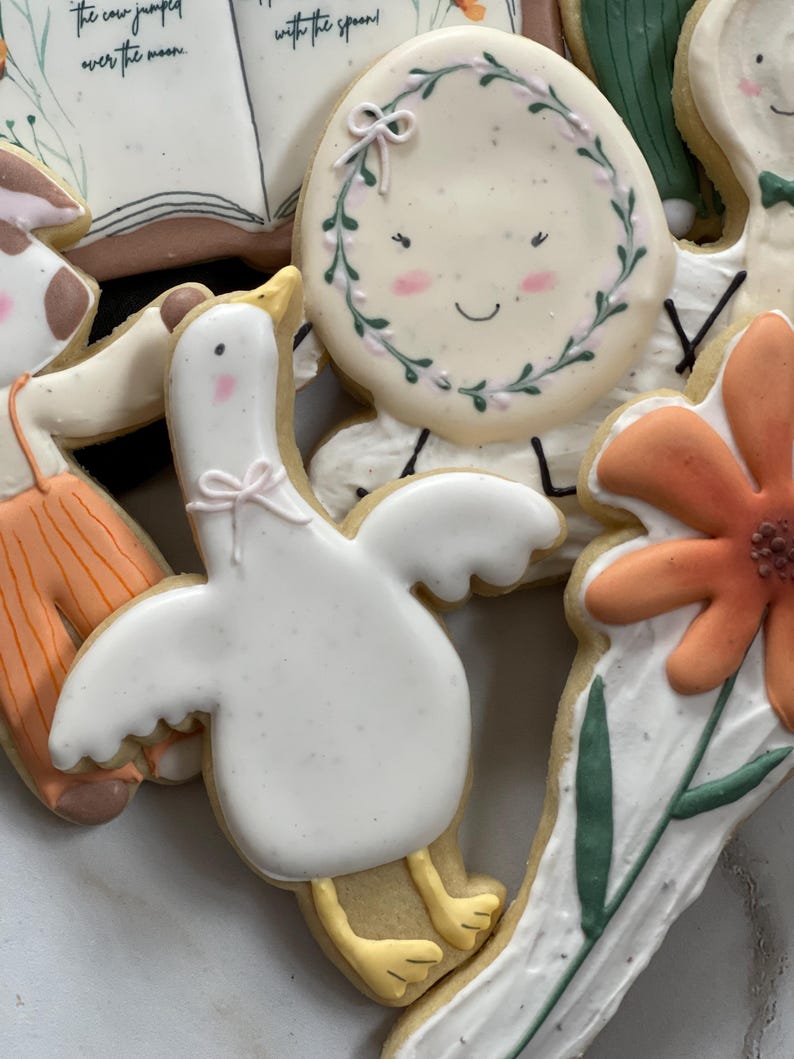 May include: A set of decorated sugar cookies featuring a white goose with yellow feet, a flower with orange petals, a book with the text "the cow jumped over the moon", and a round cookie with a wreath and a smiling face.