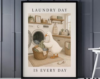 May include: A framed print depicting a laundry room with a white goose sorting laundry into a wicker basket. The text "LAUNDRY DAY IS EVERY DAY" is displayed above and below the illustration. The background includes a washing machine, shelves, and a sink.