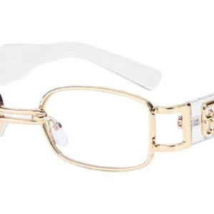 May include: Clear frame glasses with gold metal accents. The glasses have a rectangular shape and a clear plastic frame.