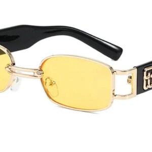 May include: Black and gold rectangular sunglasses with yellow lenses. The sunglasses have a logo on the side.