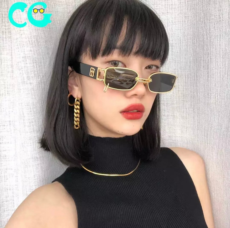May include: A woman with black hair and bangs wearing black sunglasses with a gold frame and a gold chain detail. She is wearing a black turtleneck shirt and gold earrings.