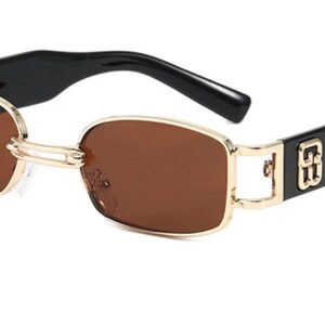 May include: Gold-rimmed sunglasses with brown lenses and black frames. The frames have a gold logo on the side.
