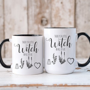 May include: Two white ceramic mugs with black handles and black rims. The mugs have a black and white graphic design with the text "There is a little Witch in all of us" and a small illustration of a salt shaker, herbs, and a heart.