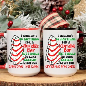 May include: Two white ceramic mugs with red and black rims. Both mugs feature a Christmas tree design with red and white stripes and green sprinkles. The text on the mugs reads "I wouldn't do anything for a Klondike Bar but I would do some sketchy stuff for Christmas Tree Cakes."