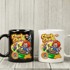 May include: Two ceramic coffee mugs with black and white handles. The mugs feature a cartoon image of a character from the horror movie Child's Play, holding a bowl of cereal. The text on the mugs reads "Chucky Charms" and "They're Magically Delicious!"