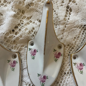 May include: Set of three white porcelain wall hooks with a gold trim and a pink rose design. Each hook has a unique shape and features a floral pattern with green leaves. The hooks are designed for wall mounting.