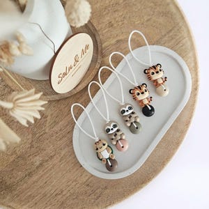 Könnte beinhalten: Vier Schnullerclips mit Tiermotiven: ein Tiger, ein Faultier und eine Schildkröte. Jeder Clip hat eine kleine Tierfigur, die an einer weißen Kordel befestigt ist. Die Clips sind auf einem weißen Tablett angeordnet, mit einem Holzschild mit der Aufschrift "Solu & Me".