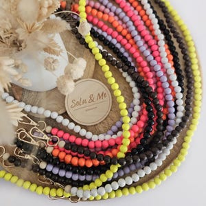 Könnte beinhalten: Eine Sammlung farbenfroher Perlen-Handy-Bänder in Gelb, Weiß, Pink, Orange, Schwarz und Lila. Die Bänder sind auf einem Holztablett mit goldfarbenen Verschlüssen und dem Text "Solu & Me accessories" angeordnet.