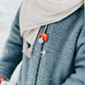 Könnte beinhalten: Nahaufnahme eines blauen Kindercoats mit einem roten und orangefarbenen Feuerwehrauto-Reißverschlussanhänger. Der Mantel wird mit einem hellbeigen Schal kombiniert. Der Reißverschlussanhänger ist ein kleines, detailliertes Spielzeug.