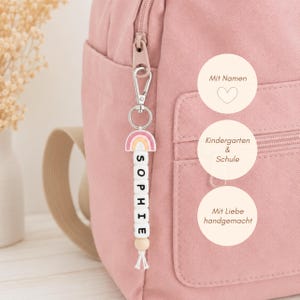 Könnte beinhalten: Ein rosa Rucksack mit einem hängenden Schlüsselanhänger, der einen Regenbogen-Anhänger und weiße quadratische Perlen mit dem Namen SOPHIE zeigt. Der Anhänger hat einen silbernen Karabinerhaken und eine Holzperle mit einer weißen Kordelquaste.