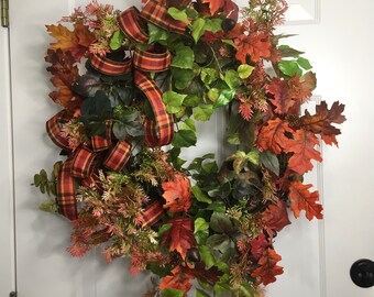 May include: A fall-themed wreath with a red, orange, and green plaid ribbon. The wreath is made of artificial leaves and branches, including oak leaves, maple leaves, and pine needles.