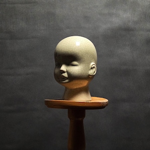 Pode incluir: Uma cabeça de boneca de cerâmica verde clara, com os olhos fechados, repousa sobre um pequeno suporte redondo de madeira. A cabeça tem um acabamento craquelado. O suporte é sustentado por um único poste de madeira torneada, sobre um fundo cinza escuro.
