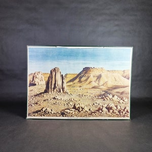 May include: A framed landscape print depicting a desert scene with towering rock formations and a vast, arid landscape under a blue sky. The print has a green border and features text at the bottom.