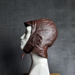 Può includere: Un casco da aviatore in pelle marrone con una piccola visiera e paraorecchie. Il casco ha una tasca con bottoni sul lato e un sottogola. La pelle appare usurata, conferendogli un'estetica vintage. Il casco è esposto su una testa di manichino bianca.