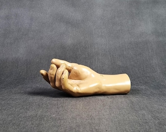 May include: A tan-coloured, life-sized hand mannequin with a slightly open palm. The fingers are slightly curved, and the skin has a smooth, glossy finish. The hand is resting on a grey surface, and the background is a solid grey.