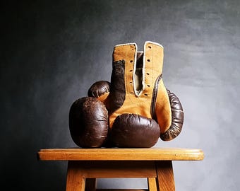 May include: A pair of vintage brown and tan leather boxing gloves on a wooden stool. The gloves have a lace-up closure and are worn.