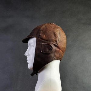 Può includere: Cappello da aviatore in pelle marrone vintage, con paraorecchie e piccola visiera. Presenta una tasca con bottone e un sottogola. La pelle mostra segni di usura, conferendogli un aspetto classico e invecchiato. Un accessorio vintage.