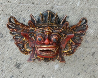 Può includere: Una maschera di legno rossa, nera e dorata con intagli intricati e occhi grandi. La maschera ha un'espressione feroce e sembra essere una maschera tradizionale balinese.
