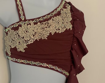 May include: A maroon dance costume with a one-shoulder design. The bodice features a layer of white lace and rhinestones. The skirt is made of a flowing fabric with a ruffled hem.