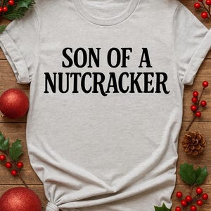 May include: A light gray t-shirt with the text "SON OF A NUTCRACKER" in black. The shirt is decorated with red Christmas ornaments, pine cones, and holly on a wooden background. A festive holiday theme.
