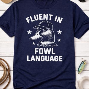 Könnte beinhalten: Marineblaues T-Shirt mit dem Text "FLUENT IN FOWL LANGUAGE" und einer Ente mit Baseballkappe. Das Design enthält Sterne. Eine beige Baseballkappe und Sonnenbrille sind im Hintergrund zu sehen. Angeltackle ist ebenfalls sichtbar.