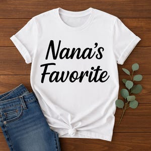 Könnte beinhalten: Weißes T-Shirt mit dem Text "Nana's Favorite" in schwarzer Schrift. Das Shirt ist an der Taille geknotet und wird mit einer blauen Jeans kombiniert. Ein Zweig mit grünen Blättern befindet sich auf der rechten Seite des Shirts.