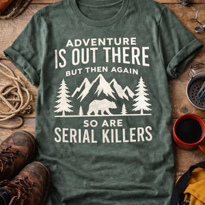 May include: Green t-shirt with the text "ADVENTURE IS OUT THERE BUT THEN AGAIN SO ARE SERIAL KILLERS" in white. The graphic includes mountains, trees, and a bear. Hiking boots, a compass, and a backpack are visible.
