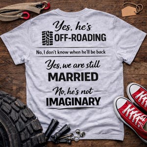 Op de afbeelding: Een lichtgrijs t-shirt met zwarte tekst met de tekst "Yes, he's OFF-ROADING. Yes, we are still MARRIED. No, he's not IMAGINARY." Rode sneakers zijn rechts zichtbaar.