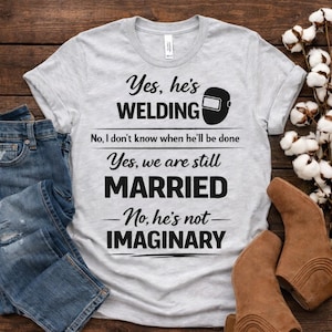 Peut inclure: Un t-shirt gris clair avec le texte "Yes, he's WELDING" et "Yes, we are still MARRIED" en noir. Un graphique de masque de soudage est au-dessus du mot "WELDING". Le t-shirt contient également le texte "No, I don't know when he'll be done" et "No, he's not IMAGINARY".
