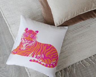 May include: White square pillow with a vibrant orange and pink tiger design. The tiger is lying down, with its stripes and facial features clearly visible. The pillow is on a textured white rug, with other pillows in the background.