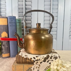 May include: A polished brass tea kettle with a curved spout, a rounded body, and a hinged metal handle. The kettle features a matching lid with a small central knob. It is positioned atop a stack of vintage books and a white lace doily.
