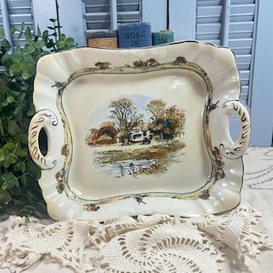 May include: Cream-colored, scalloped-edge serving tray with a landscape painting in the center. The tray has two handles and a gold trim. The painting depicts a house, trees, and a body of water.