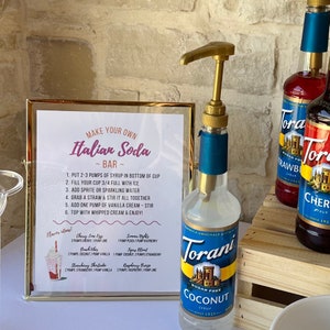 Op de afbeelding: Een gouden lijst met instructies voor het maken van Italiaanse sodas. Het bord vermeldt ingredi&euml;nten en smaakcombinaties. Op de voorgrond staat een doorzichtige fles met een gouden pompdispenser. De fles is gelabeld met "Torani Suikervrije Kokosnootsiroop Sinds 1925".