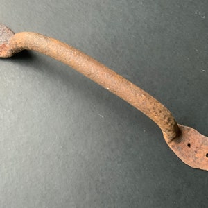 Puede incluir: Una manija de puerta de metal oxidada con forma curva y dos agujeros en cada extremo. La manija es de metal marrón oscuro con textura rugosa.