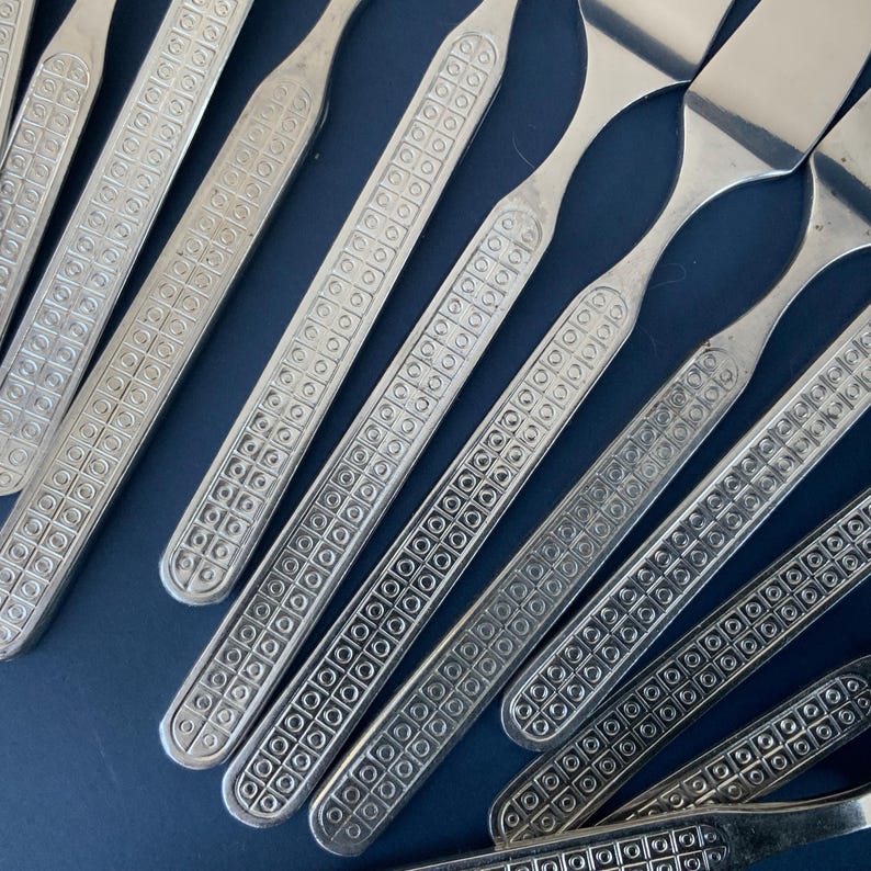 May include: Close-up of a set of silver-colored flatware. The handles have a repeating square and circle pattern. The flatware is arranged diagonally on a dark blue surface.
