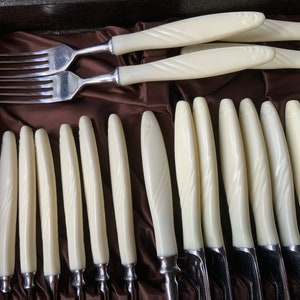 May include: A set of vintage cutlery with white plastic handles. The handles have a decorative pattern and the cutlery is arranged in a brown velvet lined box.