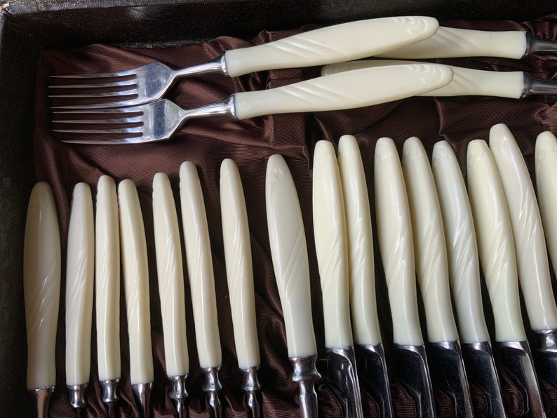 May include: A set of vintage cutlery with white plastic handles. The handles have a decorative pattern and the cutlery is arranged in a brown velvet lined box.