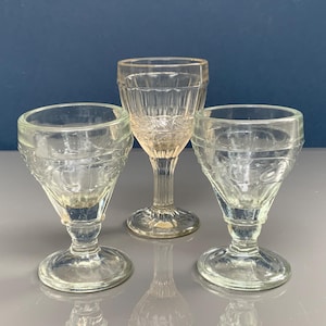 Puede incluir: Tres antiguas copas de vidrio transparente para lavado de ojos. Dos copas tienen forma cónica con base ancha y patrón detallado. La tercera copa es más alta con tallo y cuenco cilíndrico. Las tres son de vidrio transparente.