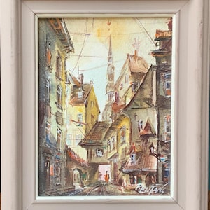 Op de afbeelding: Een aquarel schilderij van een smalle straat in een Europese stad. De gebouwen zijn hoog en smal, met kleurrijke gevels. De straat is geplaveid en er lopen mensen op. Het schilderij is onderaan rechts gesigneerd met "Reutan".