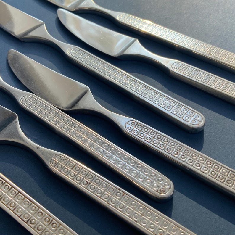 May include: Close-up of stainless steel silverware set, including knives and forks. The handles feature a repeating square pattern. The silverware is arranged diagonally on a dark blue surface, with the blades of the knives reflecting light.