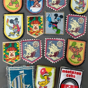 Op de afbeelding: Een verzameling vintage stoffen patches met diverse ontwerpen. De patches tonen stripfiguren, schepen, paddenstoelen en tekst in meerdere talen. De patches hebben een decoratieve rand en zijn in verschillende kleuren, waaronder blauw, geel en rood.