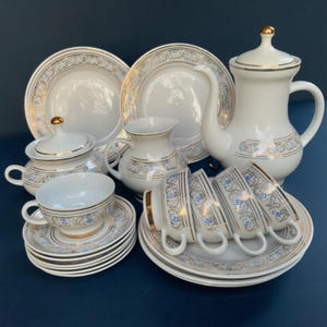 Könnte beinhalten: Vintage weißes Porzellan-Teeservice mit goldenen Verzierungen und blauen Blumenmustern. Das Set umfasst eine Kaffeekanne, eine Zuckerdose, einen Sahnekännchen, Tassen, Untertassen und Teller. Das Teeservice steht auf einer dunkelblauen Oberfläche.