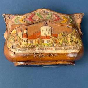 May include: A brown, wooden decorative box featuring a relief of a castle with red-tiled roofs, towers, and surrounding trees. The box is labeled "Riga - Pils" and has decorative accents in red, yellow, and green.