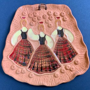 May include: A terracotta-colored ceramic wall plaque featuring three figures in relief. Each figure wears a black bodice, light-colored sleeves, and a red plaid skirt. The background has a textured, straw-like pattern with small circular accents.