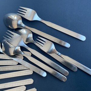 May include: A collection of stainless steel silverware, including forks and spoons, arranged on a dark blue surface. The silverware has a simple, modern design with long, slender handles. Some pieces have the brand name stamped on them.
