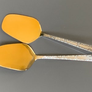 Puede incluir: Dos cucharas de servir de color dorado con mangos de plata adornados. Las cucharas tienen una forma redondeada ligeramente curvada.