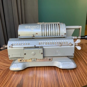 Puede incluir: Una calculadora mecánica vintage de color plateado con el texto "Феликс М" en la parte superior. La calculadora tiene una serie de diales numerados y una palanca en el lateral.