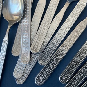 May include: Close-up of a collection of silver-colored flatware, including spoons and forks. The handles feature a repeating square and circle pattern. The flatware is arranged on a dark blue surface.