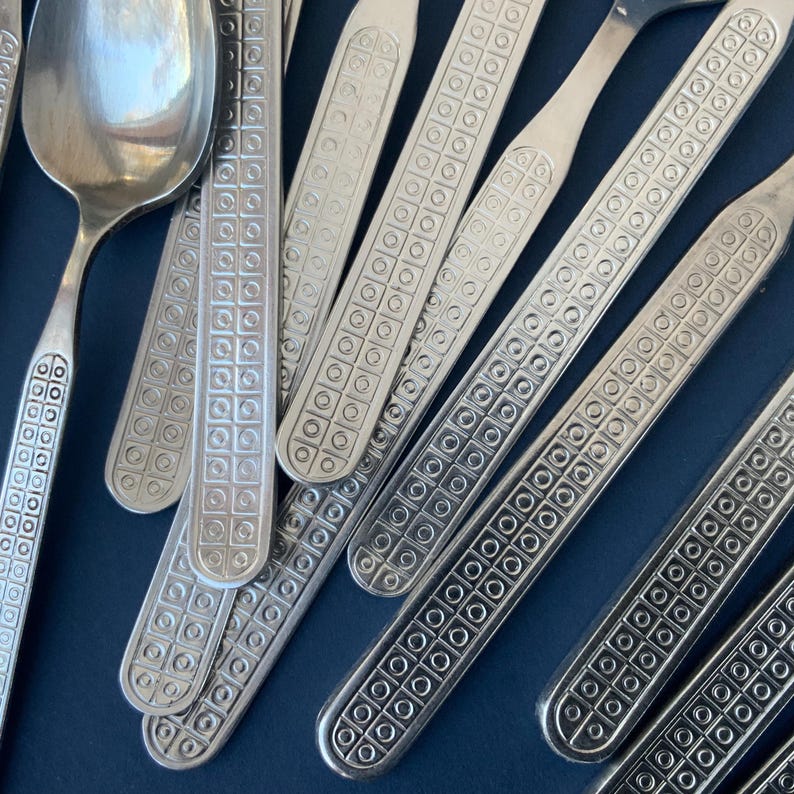 May include: Close-up of a collection of silver-colored flatware, including spoons and forks. The handles feature a repeating square and circle pattern. The flatware is arranged on a dark blue surface.