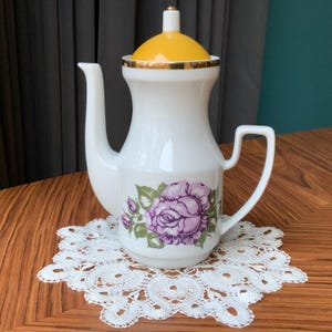 Könnte beinhalten: Weiße Teekanne aus Keramik mit gelbem Deckel und einem Blumendesign mit violetten Rosen. Die Teekanne hat einen goldenen Rand um den Deckel und die Basis.
